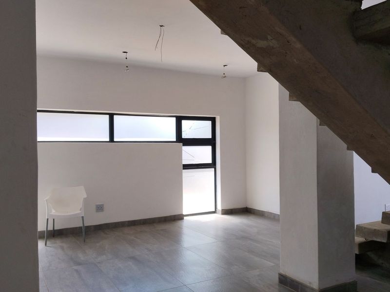 An open-plan living area featuring modern grey floor tiles, aluminium-framed windows, and an unfinished concrete staircase, awaiting final interior finishes.