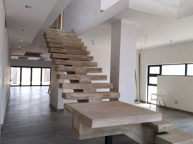 Spacious open-plan living area featuring a striking raw concrete staircase, large windows, and tiled flooring, currently in an unfinished state awaiting final touches.