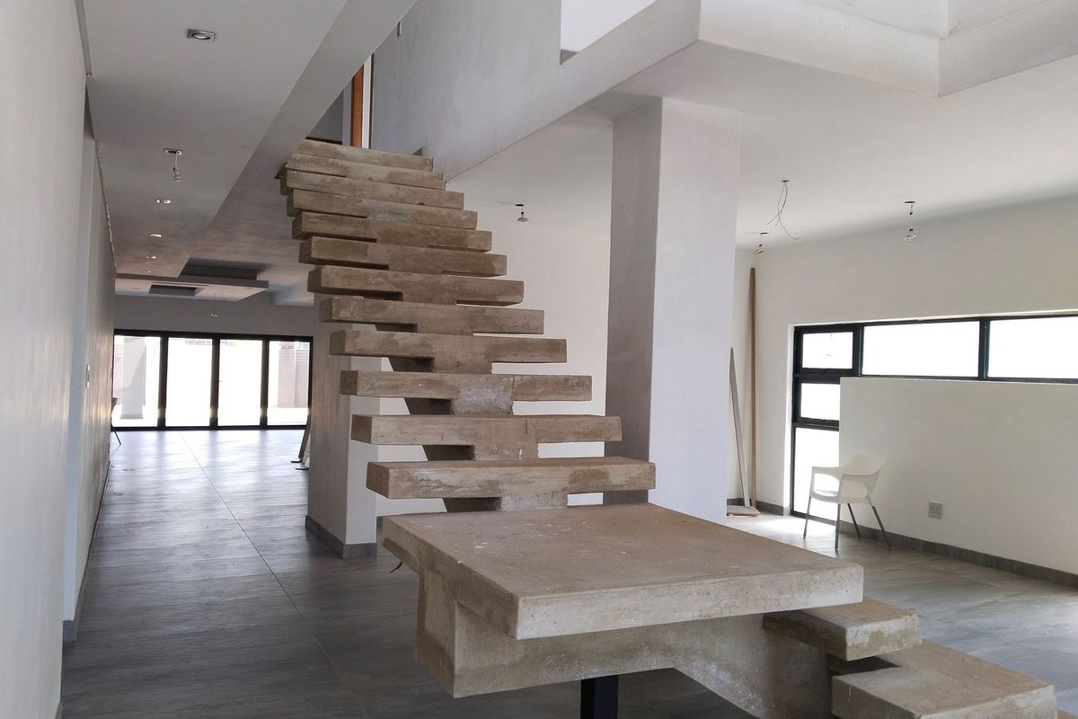 Spacious open-plan living area featuring a striking raw concrete staircase, large windows, and tiled flooring, currently in an unfinished state awaiting final touches.