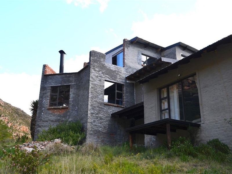 Rear view of a multi-level farm property featuring exposed brickwork, a metal chimney, and a timber deck, currently in a state of significant disrepair.