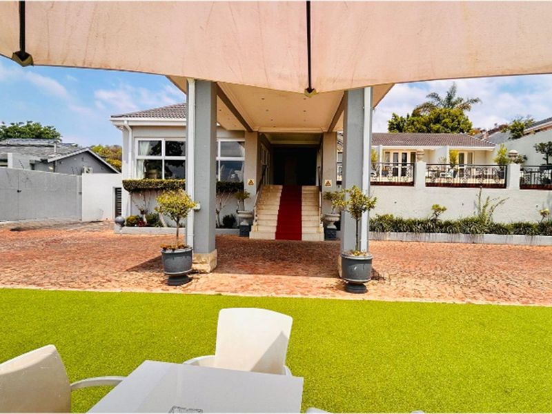 A spacious exterior entrance area featuring a covered walkway, paved driveway, and landscaped garden space leading to the main building entrance.