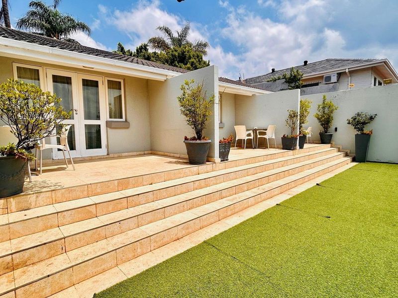 A well-maintained rear patio featuring tiled steps leading to a paved outdoor entertainment area, adjacent to a neat artificial lawn.