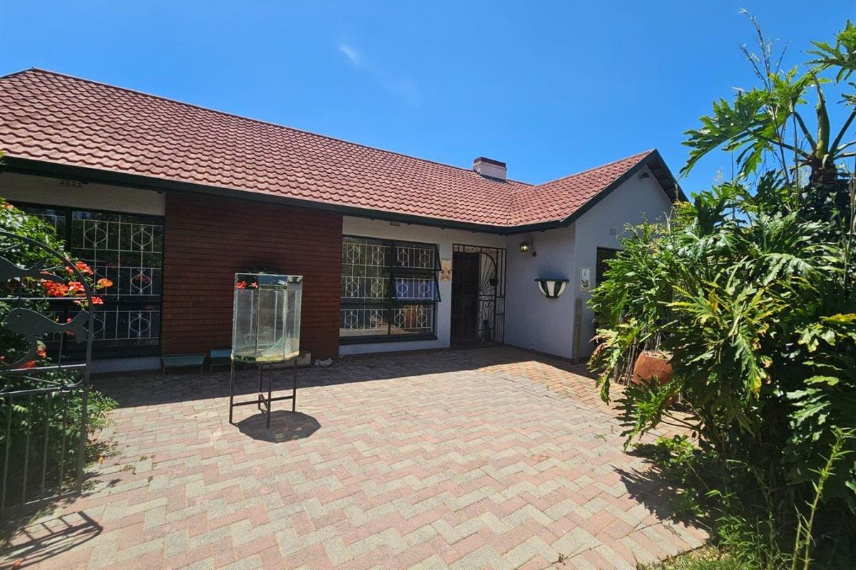 A rear exterior view of the house featuring a paved patio area, brick facade, and tiled roof with security features on windows and doors.