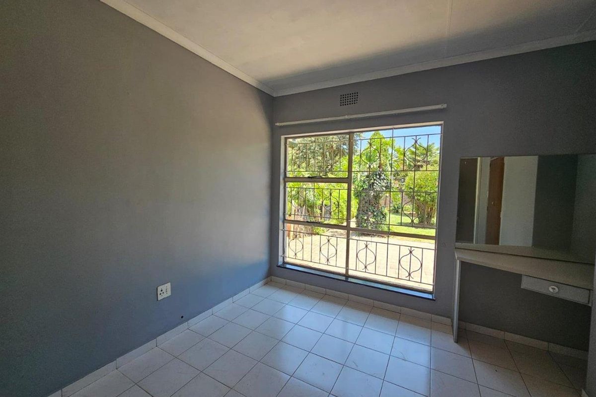 A bright, empty bedroom featuring tiled flooring, a built-in vanity, and large windows fitted with security bars overlooking the garden.
