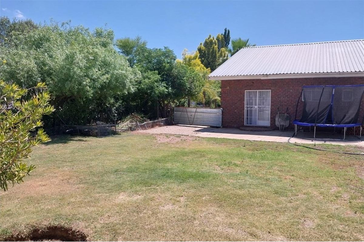 A spacious rear garden featuring a paved patio area, established trees, and a brick exterior facade with a metal roof.