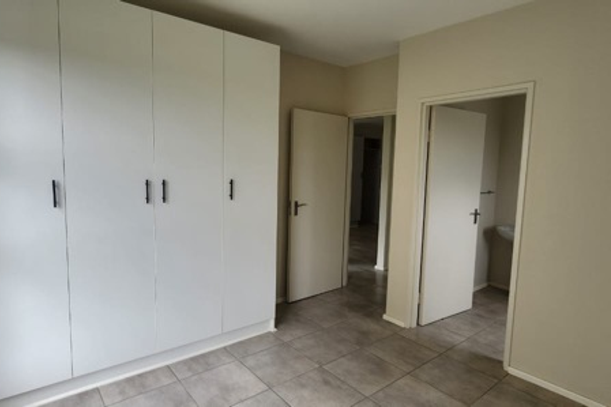 A clean, empty bedroom featuring neutral tiled flooring and ample built-in cupboard storage, with direct access to an adjoining room.
