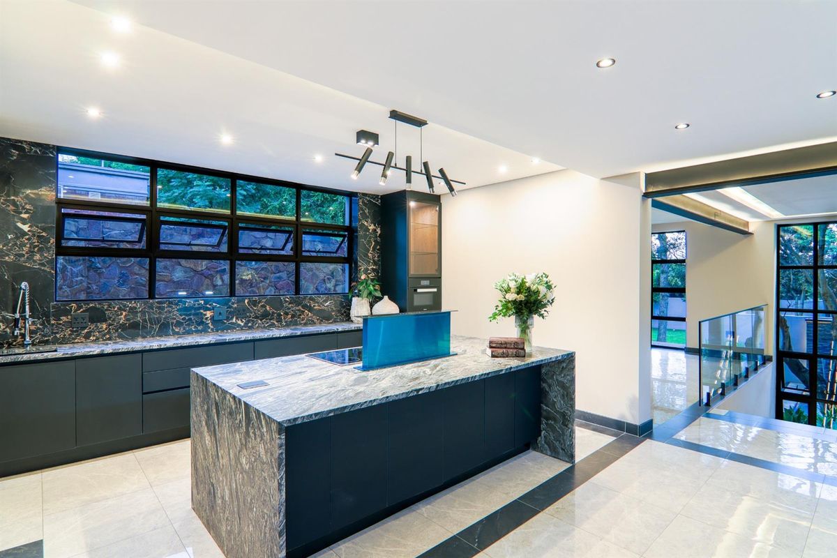 Modern kitchen featuring a large stone-topped island, integrated hob with pop-up extractor, dark cabinetry, and high-end finishes with large windows.