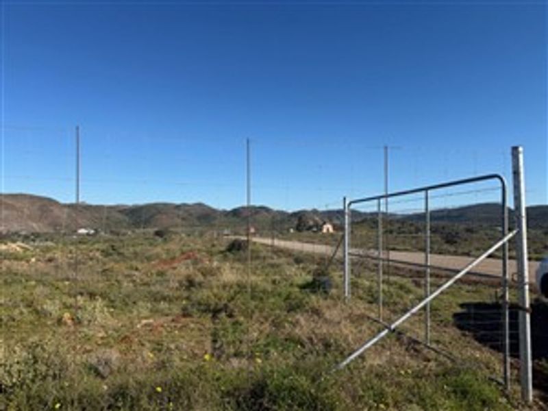 A view of the vacant residential land featuring wire perimeter fencing, a metal access gate, and frontage onto a gravel road.