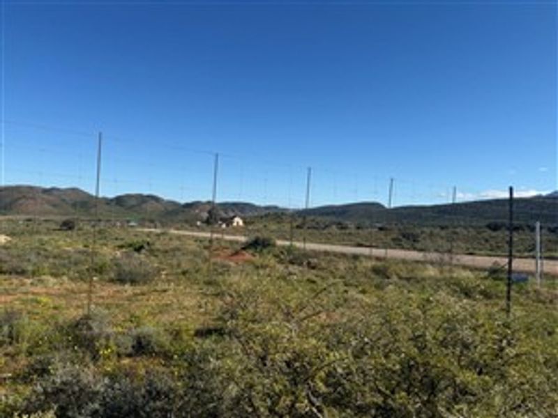 A view of a vacant residential plot featuring wire fencing and a dirt road with mountain views in the background.