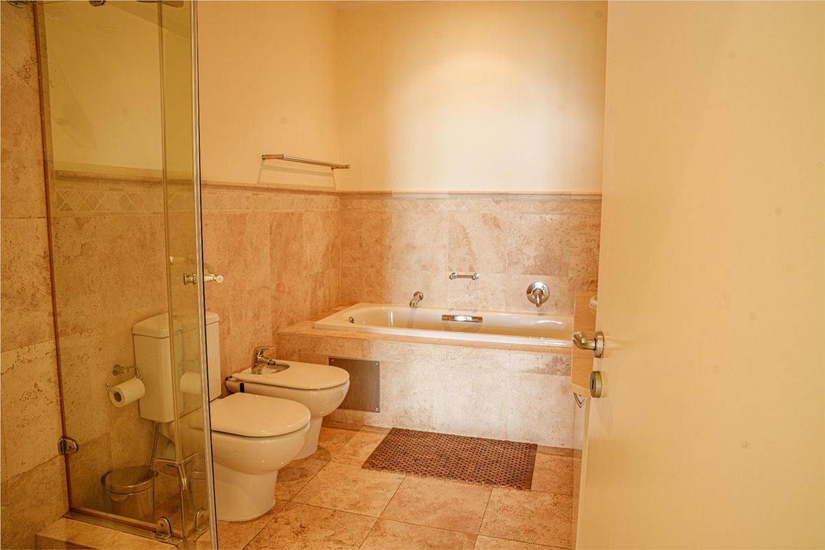 A well-maintained bathroom featuring a built-in bathtub, bidet, toilet, and a glass-enclosed shower, finished with neutral stone-look wall and floor tiles.