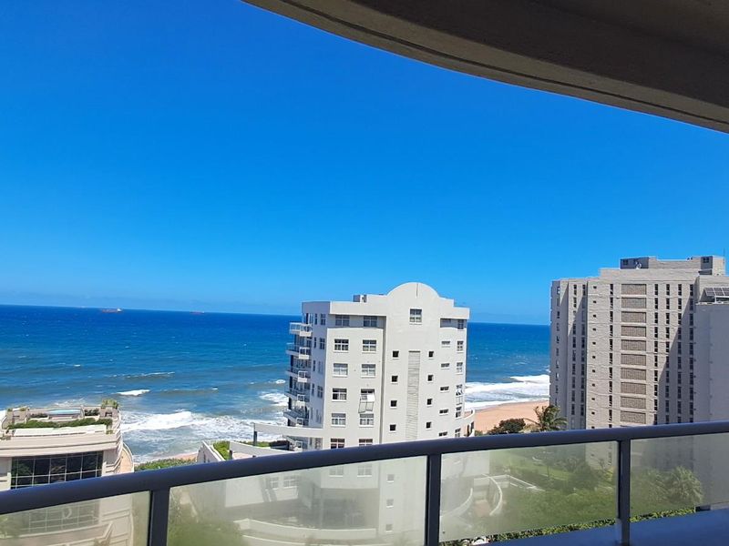 A clear, elevated ocean view captured from a balcony with a modern glass balustrade, overlooking surrounding coastal apartment buildings.