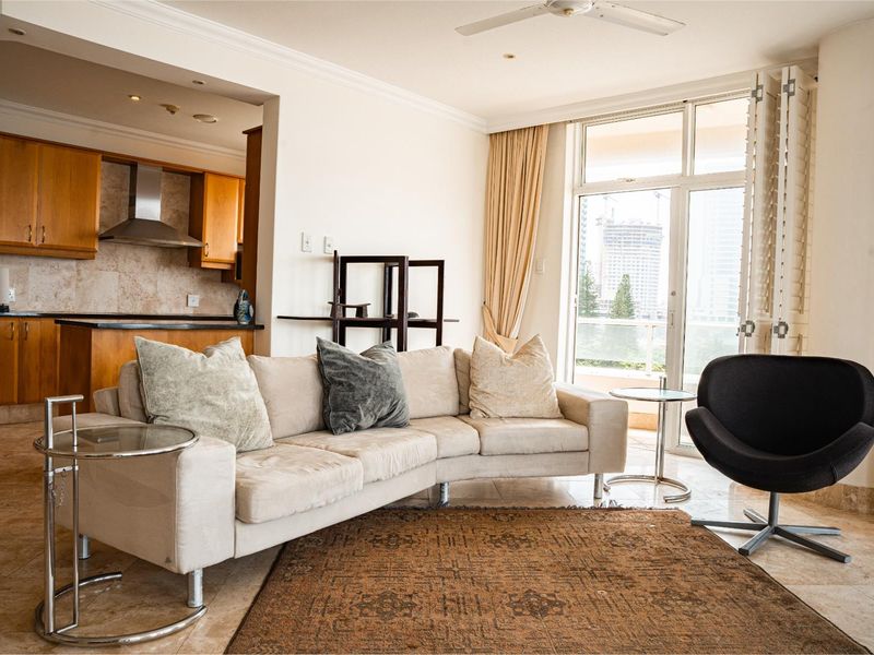 Open-plan living area featuring stone flooring, a ceiling fan, and an adjoining kitchen with wood cabinetry and stone countertops, leading to a balcony.