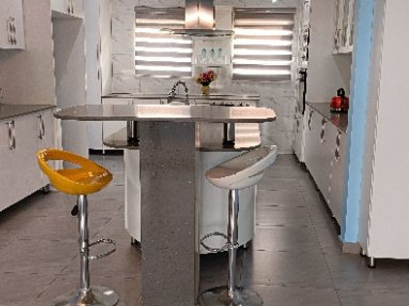 A functional kitchen featuring white cabinetry, a central island with seating, tiled flooring, and modern roller blinds.