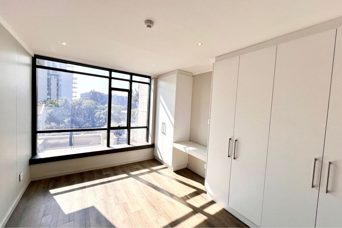 A bright, modern bedroom featuring large windows with natural light, wood-look flooring, and extensive built-in white cabinetry with an integrated desk area.