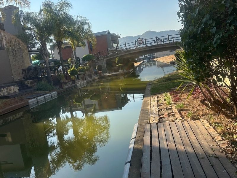 A scenic view of a residential canal featuring a pedestrian bridge, timber jetty, and landscaped waterfront surroundings.