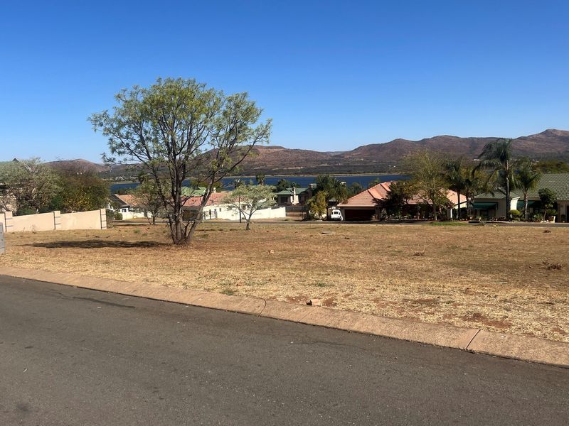A vacant residential plot featuring a gentle slope, established trees, and scenic views of the surrounding landscape and distant water.