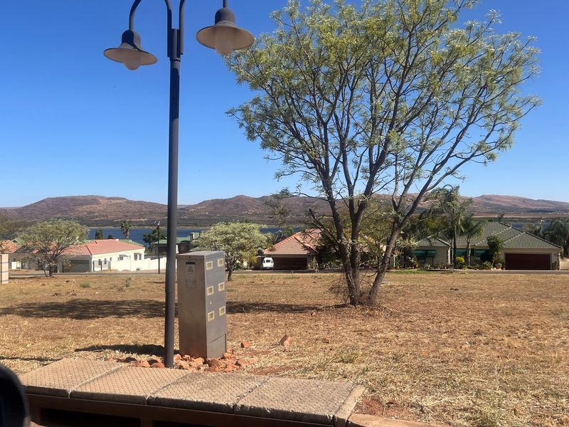 A view of a vacant residential plot featuring a street lamp, utility box, and mature tree, with surrounding neighborhood and distant mountain views.