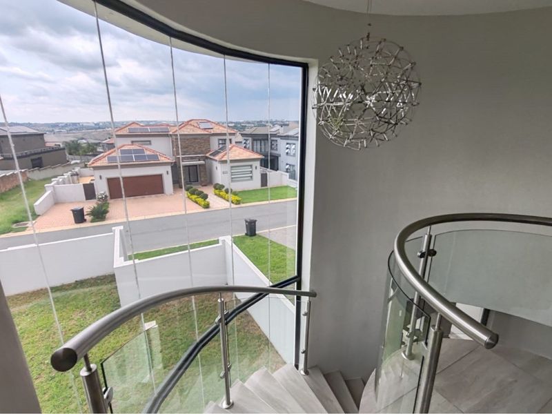 Elegant curved staircase with stainless steel balustrade and expansive floor-to-ceiling windows offering a bright, modern aesthetic and views of the surrounding estate.