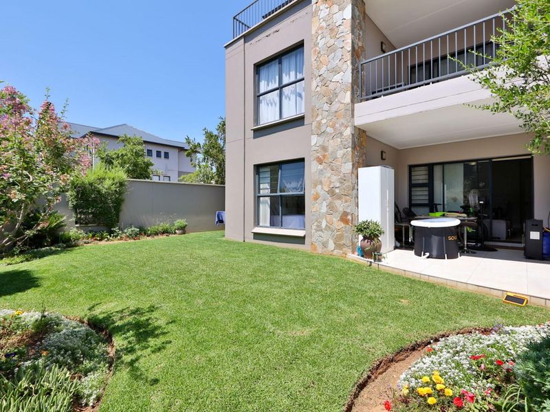 A well-maintained rear garden featuring a tiled patio, stone-clad exterior wall, and a balcony overlooking the lawn.