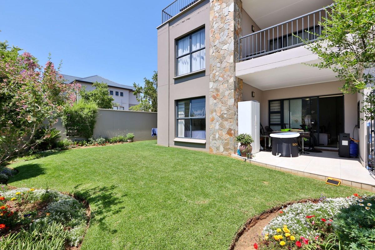 A well-maintained rear garden featuring a tiled patio, stone-clad exterior wall, and a balcony overlooking the lawn.