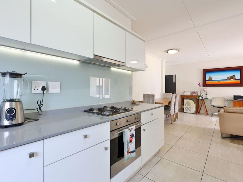 Modern open-plan kitchen featuring sleek white cabinetry, stone countertops, glass splashback, and integrated stainless steel appliances, flowing into the dining and living area.