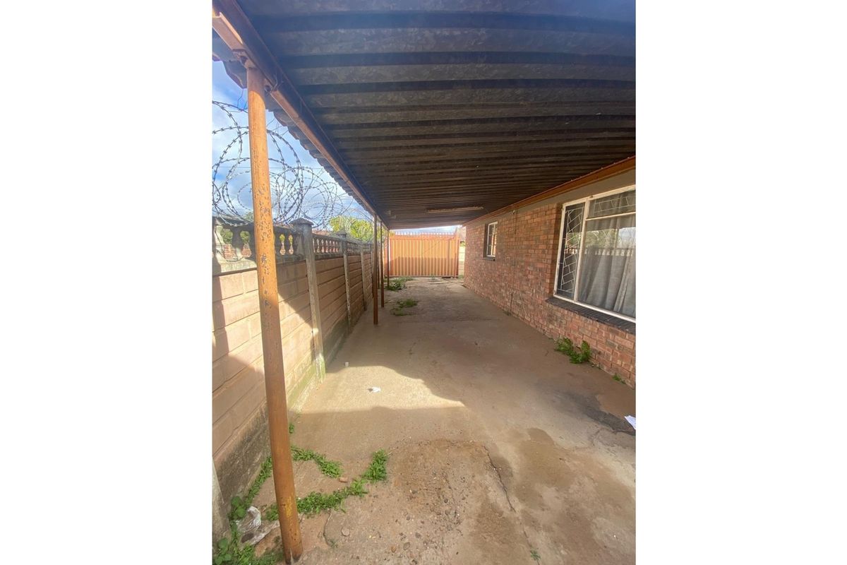 A covered outdoor walkway or carport area featuring brick walls, concrete flooring, and a metal roof structure with security fencing.