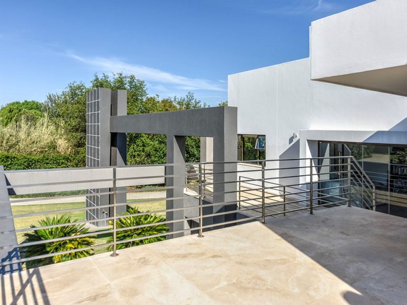 A spacious modern balcony featuring a sleek stainless steel balustrade and large format floor tiles, overlooking the property's architectural exterior and garden.