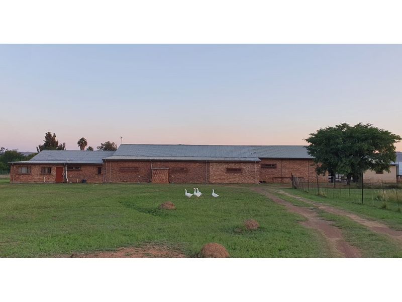 A wide exterior view of a brick farm building with a metal roof, set within a large grassy field.