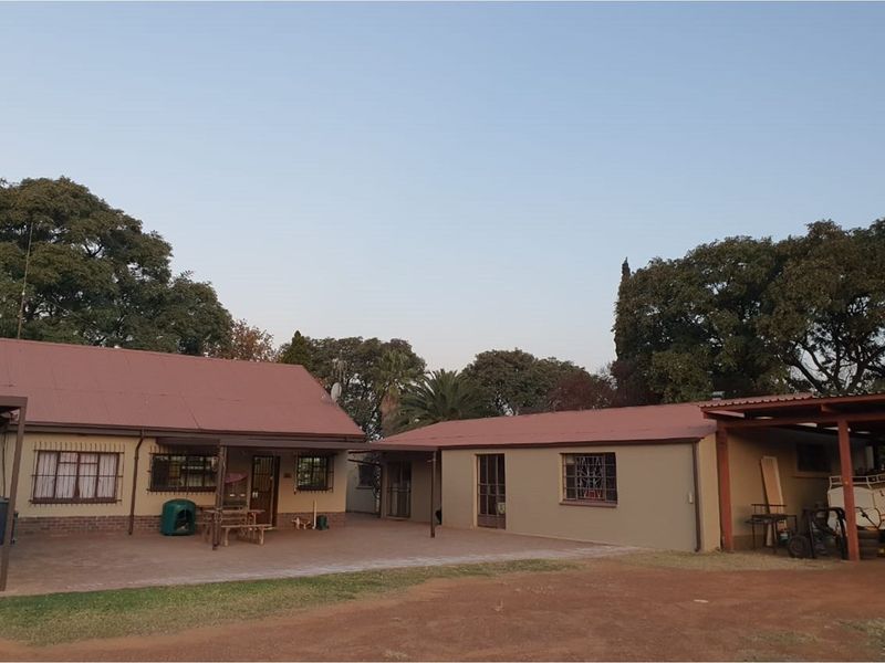 A view of the farm dwelling featuring a paved courtyard area, metal roof, and covered outdoor spaces surrounded by mature trees.