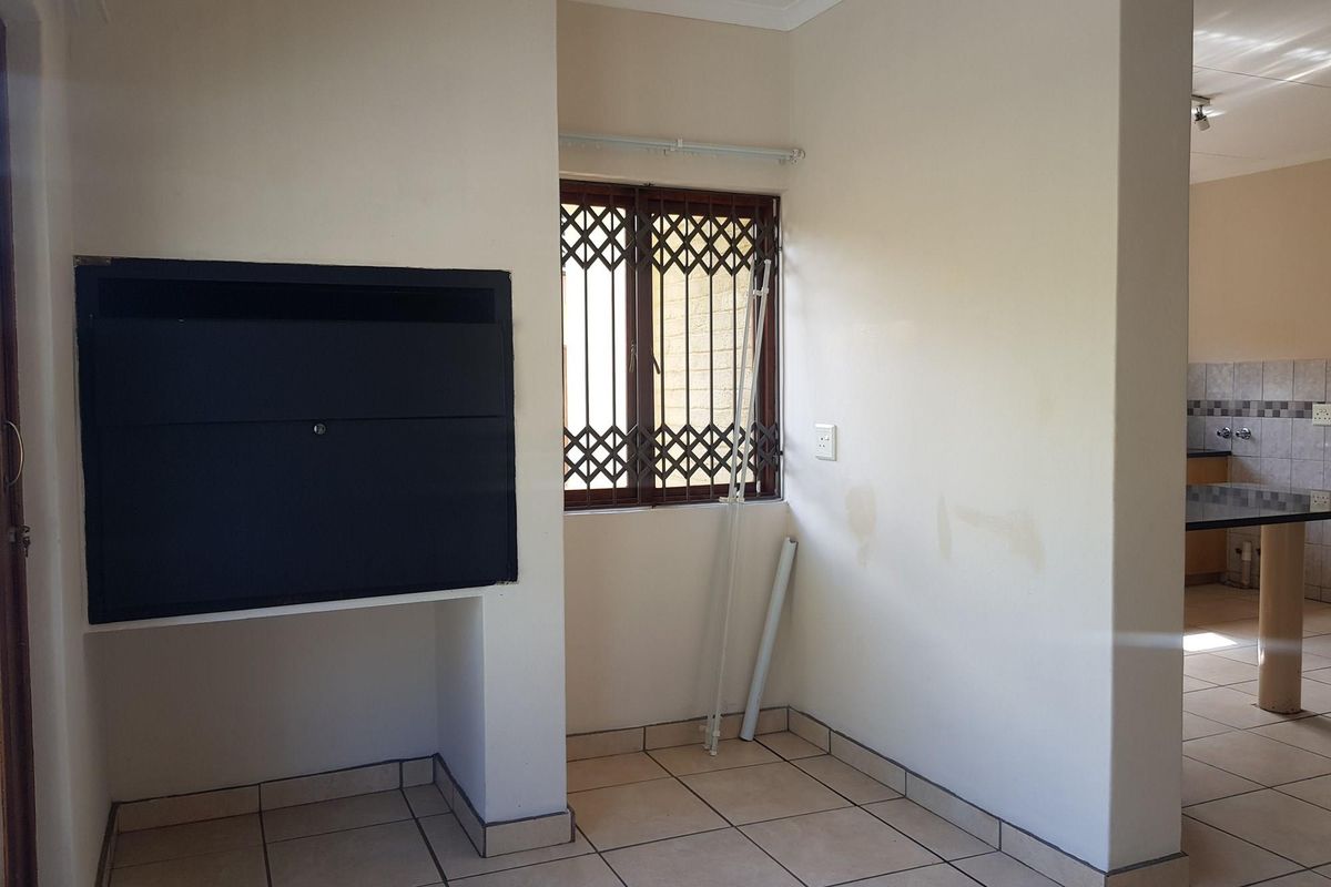 An empty living area featuring a built-in braai, tiled flooring, and a window with a security gate, leading through to the kitchen area.
