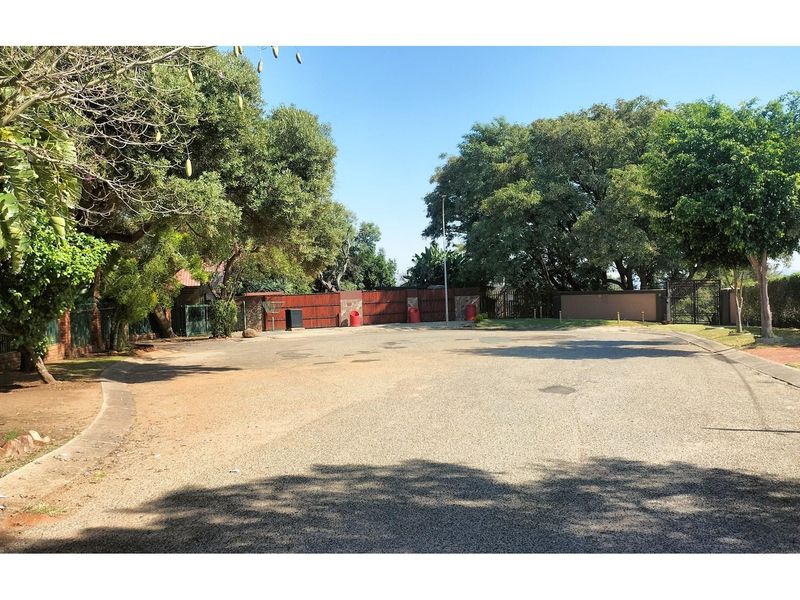 A view of the residential street approach featuring mature trees, paved road surface, and perimeter boundary fencing.