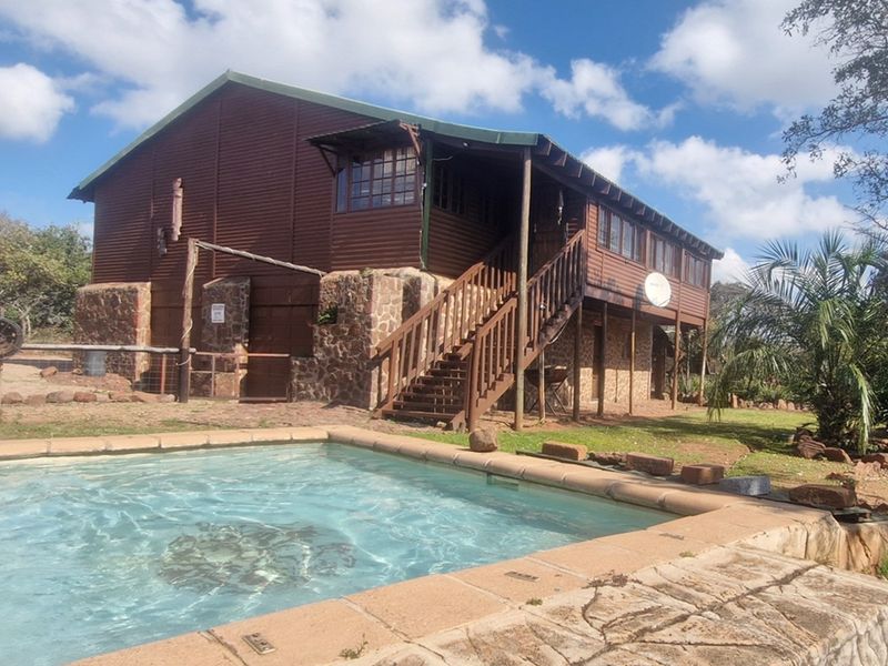 A rustic timber-clad house featuring an elevated deck, external staircase, and a private swimming pool set within a natural garden landscape.