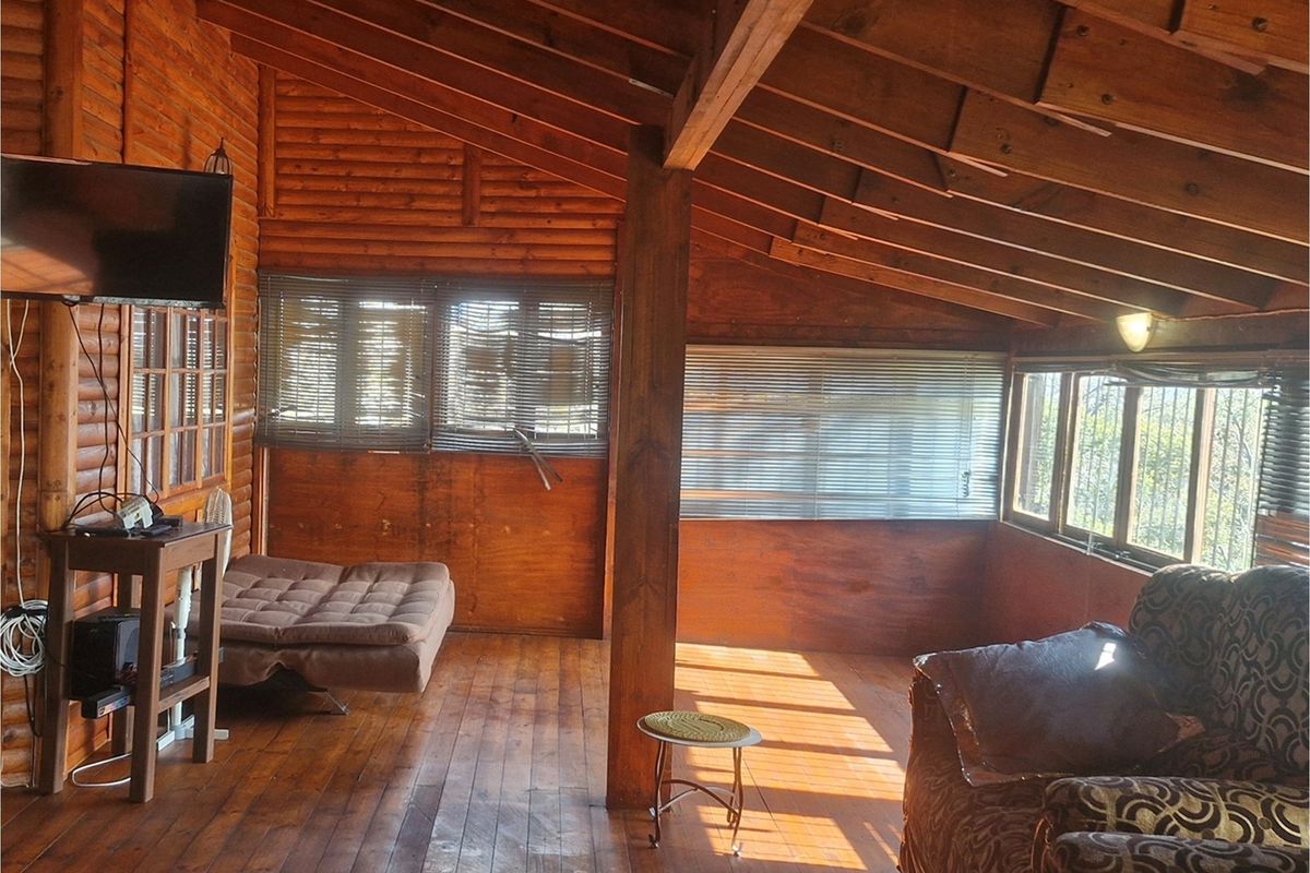 A rustic living room featuring exposed timber beam ceilings, wood-paneled walls, and large windows with fitted blinds, creating a warm, cabin-style atmosphere.