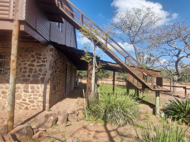 Exterior view of a rustic stone and timber house featuring a raised wooden staircase leading to an upper level and a natural garden setting.