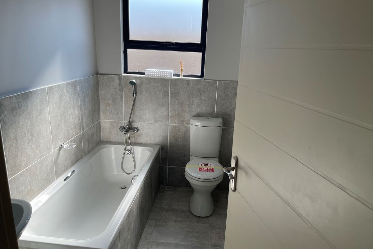 A modern bathroom featuring a built-in bathtub with a handheld shower mixer, a standard toilet, and neutral grey wall and floor tiling.