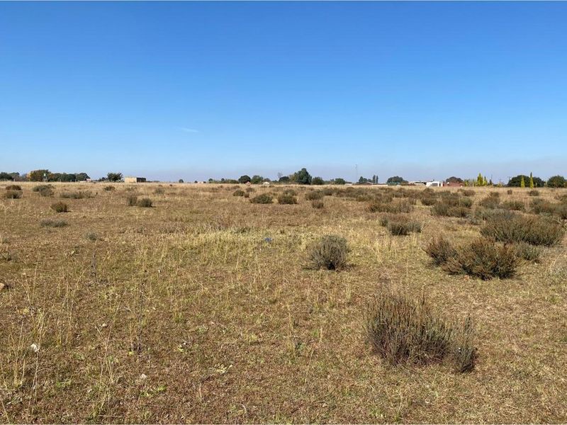 A wide-angle view of a vacant residential plot featuring natural dry grass and low-lying shrubs under a clear blue sky.
