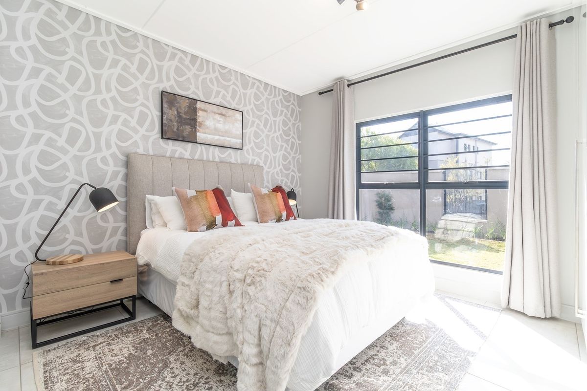 A well-lit bedroom featuring a patterned accent wall, large window with security bars, and neutral tiled flooring.