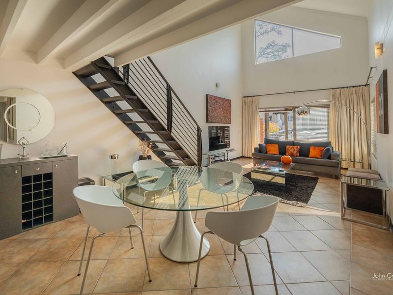 Spacious open-plan living and dining area featuring high ceilings, exposed beams, a striking metal staircase, and large windows providing abundant natural light.