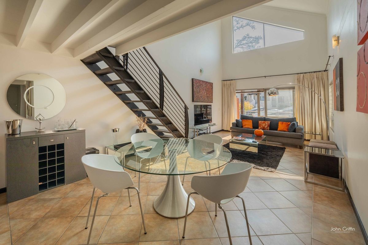 Spacious open-plan living and dining area featuring high ceilings, exposed beams, a striking metal staircase, and large windows providing abundant natural light.