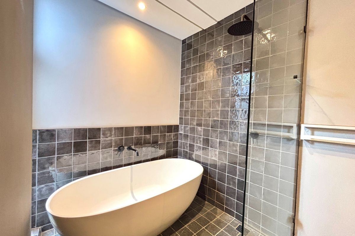 Modern bathroom featuring a freestanding bathtub, walk-in shower with glass screen, and dark tiled finishes. Requires completion of tiling around the faucet.