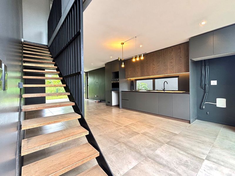 Modern open-plan kitchen and living area featuring sleek dark cabinetry, integrated appliances, tiled flooring, and a contemporary floating staircase with a metal balustrade.