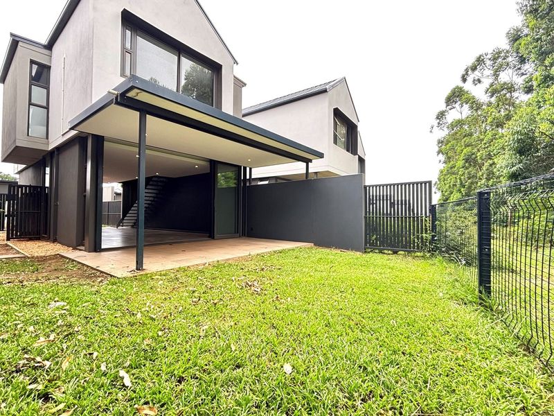 Rear view of a modern townhouse featuring a covered patio, tiled outdoor area, and a private lawn enclosed by metal fencing.