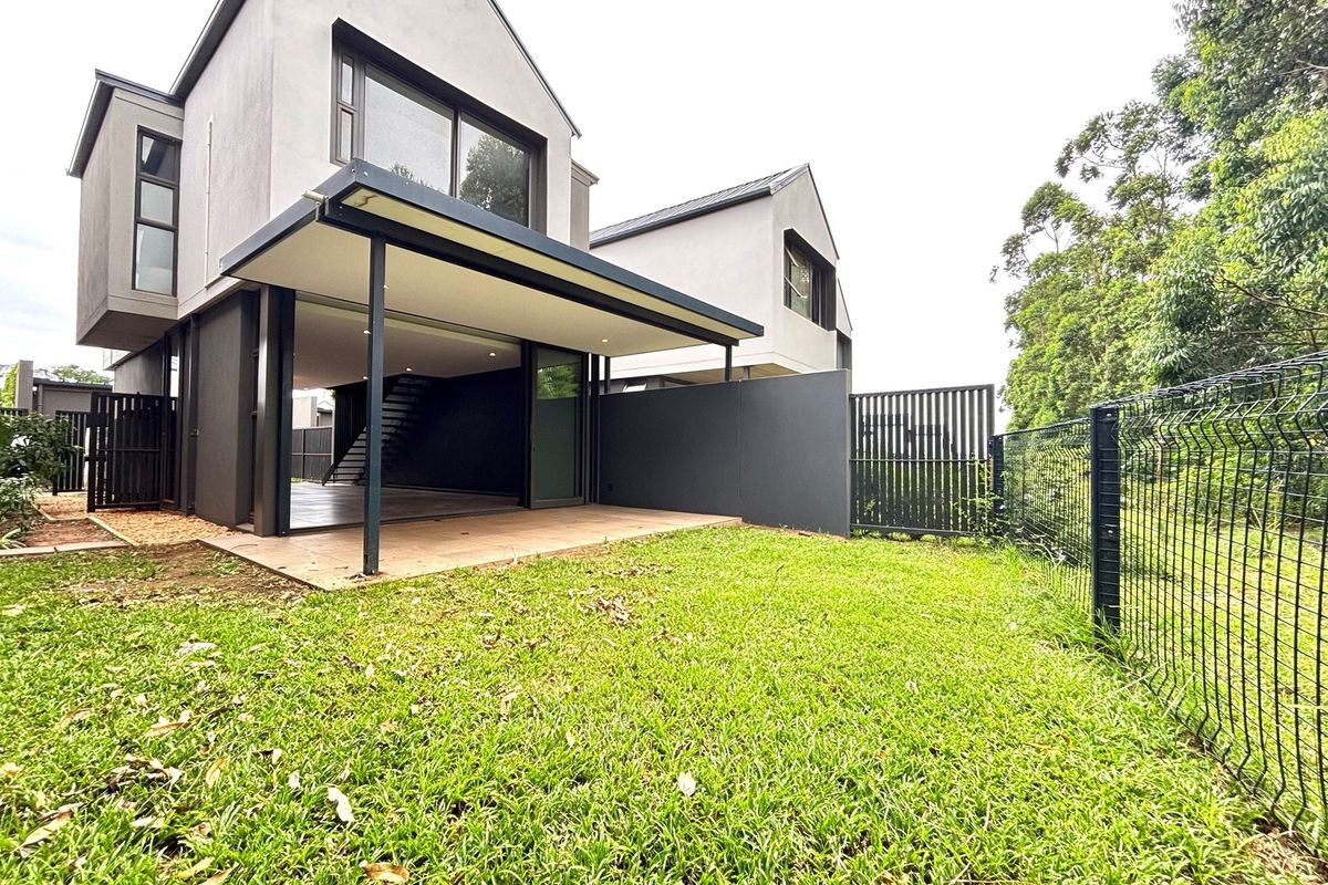 Rear view of a modern townhouse featuring a covered patio, tiled outdoor area, and a private lawn enclosed by metal fencing.