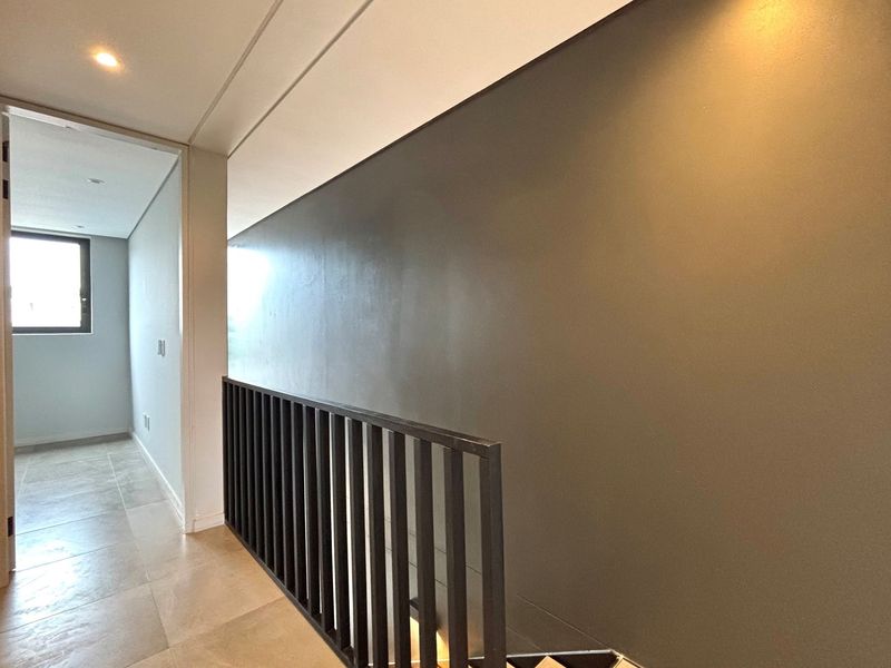 A bright and modern hallway featuring tiled flooring, a sleek metal balustrade, and recessed lighting, leading towards a sunlit room.