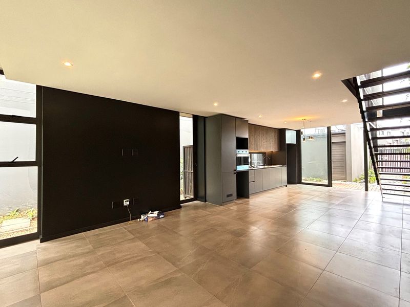 Modern open-plan living and kitchen area featuring sleek dark cabinetry, built-in oven, large format floor tiles, and a contemporary metal staircase.