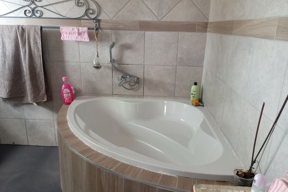 A corner bathtub featuring neutral ceramic wall tiles and a wood-look tiled surround, equipped with a wall-mounted shower mixer and handheld showerhead.
