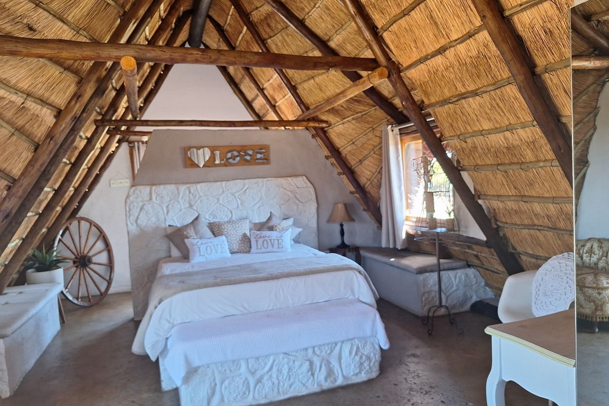 A rustic bedroom featuring a high vaulted thatched roof, exposed timber beams, and a unique stone-textured headboard wall.