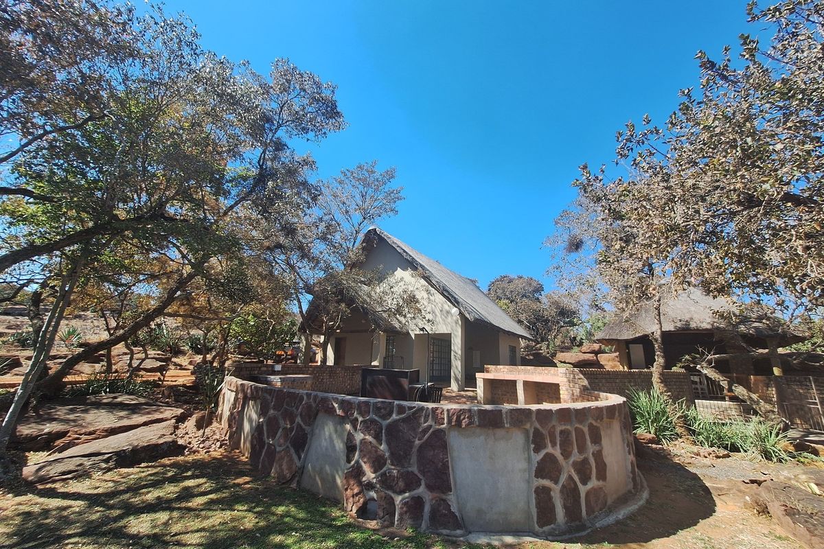 A rustic farm-style dwelling featuring a thatched roof and a curved stone-clad outdoor entertainment wall set within a natural rocky landscape.