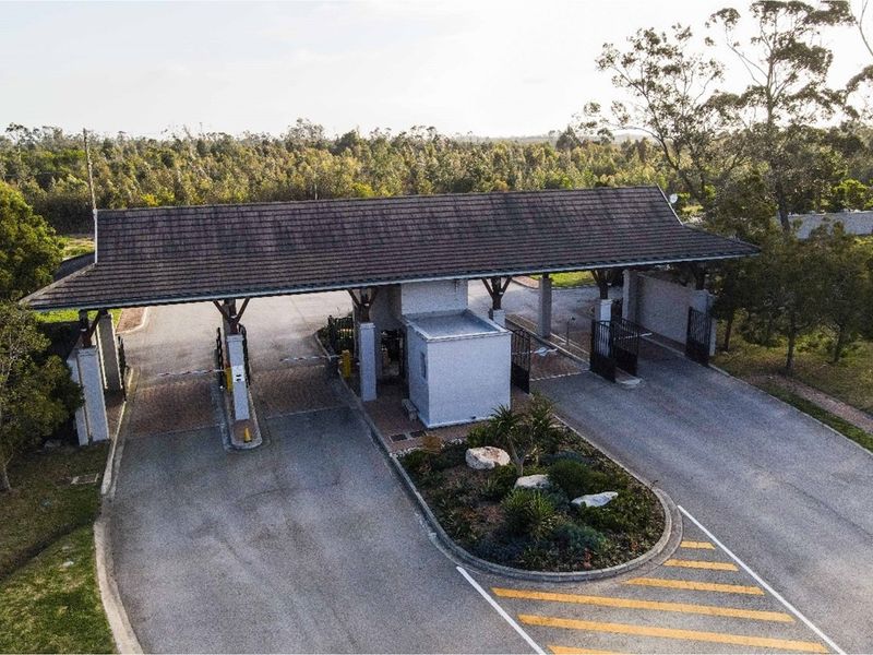 Aerial view of a secure residential estate entrance featuring a gatehouse, boom gates, and a paved driveway with a landscaped traffic island.
