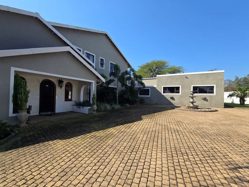 Spacious front exterior featuring a large paved driveway, a central stone fountain, and an arched entrance with established landscaping.